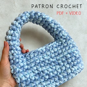 May include: A crocheted handbag in shades of light blue and white, with a rounded handle. The bag is made from thick yarn and displays the text "PATRON CROCHET PDF + VIDEO" in black and red.