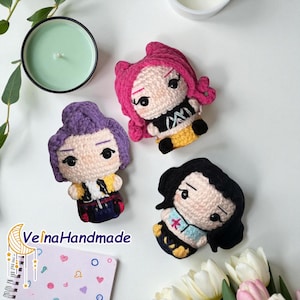 May include: Three handmade crochet dolls with pink, purple, and black hair. Each doll has a unique outfit, with yellow and black details. The dolls are arranged on a white surface, with a candle and flowers in the background.