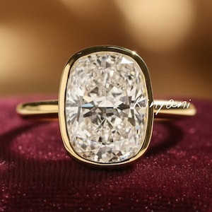 May include: A gold ring with a large, cushion-cut diamond. The diamond is set in a bezel setting, and the band is a simple, polished gold. The ring is displayed on a burgundy velvet surface.