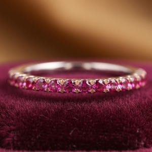 May include: A delicate ring featuring a band of vibrant pink gemstones. The ring's setting is a light metal, and the gemstones are closely set around the band. The ring is displayed on a deep red velvet surface.