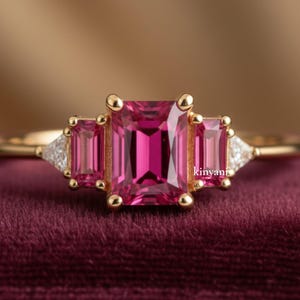 May include: A gold ring featuring a large, rectangular pink gemstone flanked by two smaller, rectangular pink gemstones. The ring has a delicate band and small, round gold accents around the gemstones. Small white accent stones are on either side of the smaller gemstones.