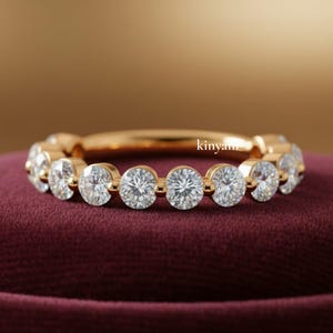 May include: A gold ring featuring a series of round, clear gemstones set around the band. The ring is displayed on a dark red surface. The word "kinyan" is visible on the ring.