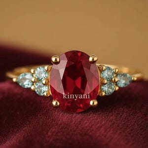 May include: A gold ring with a large, oval-cut, deep red gemstone at its centre, complemented by clusters of smaller, light blue gemstones. The ring is set against a burgundy backdrop, with the word "kinyani" visible.