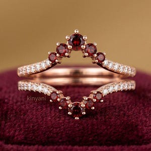 Könnte beinhalten: Ein Ring-Set aus Roségold in Chevron-Form, bestehend aus zwei V-förmigen Bändern. Die Ringe sind mit kleinen, runden, dunkelroten Granat-Edelsteinen und winzigen klaren Akzentsteinen entlang der Metallschienen besetzt. Der Text kinyani ist sichtbar.