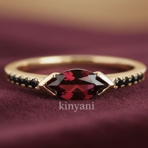 May include: A gold ring with a marquise-cut, deep red gemstone as the centerpiece. The band is adorned with small black stones. The ring is set against a burgundy background. The word "kinyani" is printed on the ring.