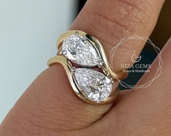 May include: A gold ring featuring two pear-shaped diamonds. The diamonds are set side-by-side, with a flowing gold band that curves around the finger. The ring is shown on a person's finger.
