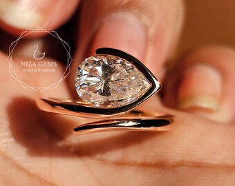 May include: A bypass ring featuring a pear-cut clear gemstone set in a polished rose gold band. The design includes a curved metal setting that partially wraps around the faceted stone, creating an open, asymmetrical silhouette on the finger.