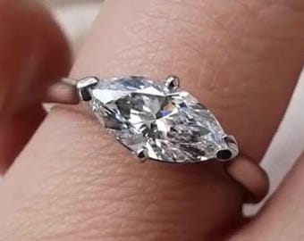 May include: A marquise-cut diamond ring with a thin, silver band. The diamond is set with four prongs and sparkles brightly. The ring is shown on a finger.