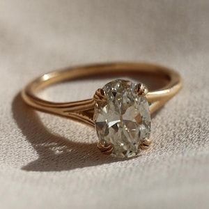 May include: A gold ring with an oval-cut diamond. The ring's band is a thin, delicate design with a split shank. The diamond is the focal point, sparkling against a neutral background. The ring is a piece of jewelry.