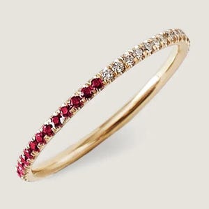 Op de afbeelding: Een gouden bandring met een half-eeuwigheidsontwerp. De ring heeft aan één kant een rij kleine, ronde, rode edelstenen en aan de andere kant heldere edelstenen. De ring staat tegen een neutrale achtergrond.