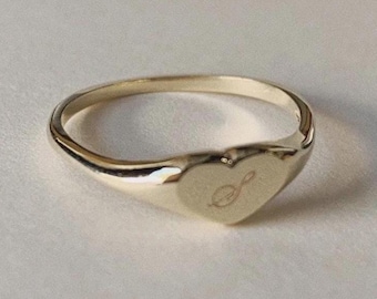 May include: A gold-colored ring with a heart-shaped face. The heart has a small, engraved design. The ring is set against a white background.