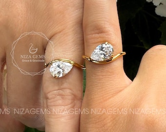 May include: Two gold rings, each featuring a pear-shaped diamond. The rings are displayed on fingers, with a backdrop of white flowers and green leaves. The rings have a delicate, minimalist design.