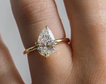 May include: A gold ring featuring a pear-shaped diamond. The diamond is set with prongs and sits atop a thin, polished gold band. The ring is shown on a person's finger.