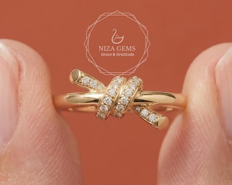 May include: A gold-colored ring with a knot design, encrusted with small, clear gemstones. The ring is held between two fingers against a warm, orange-brown background. The ring's design features a knot shape with the gemstones set along the knot's curves.