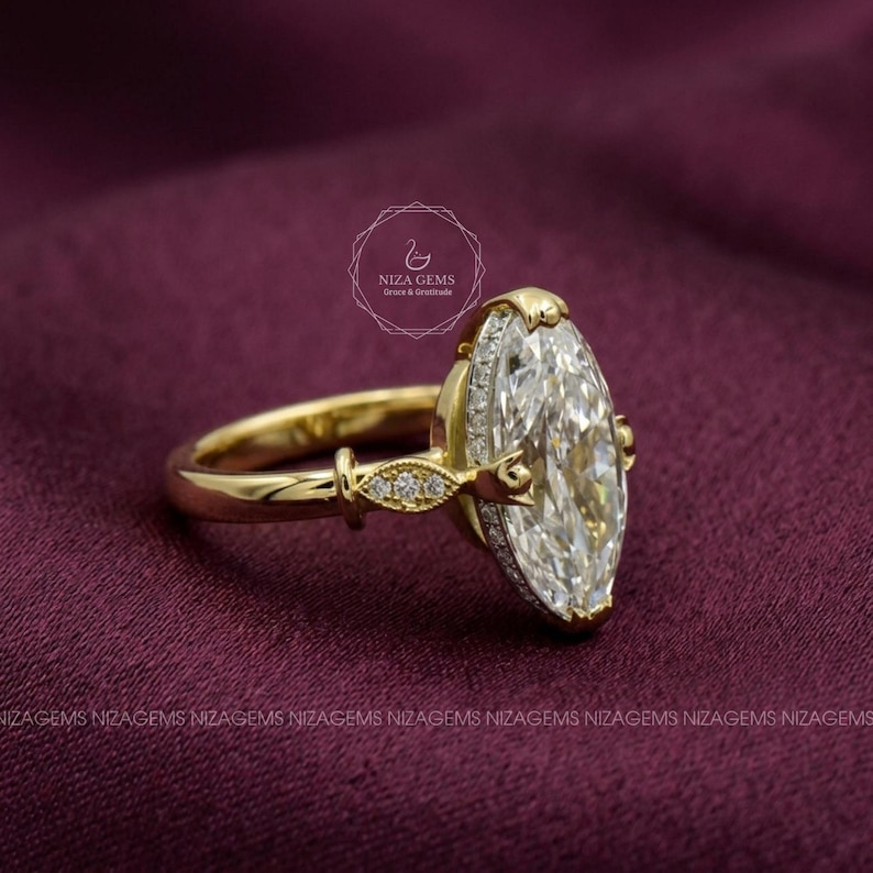 May include: A gold engagement ring with a large, marquise-cut diamond as the centerpiece. The ring features a halo of smaller diamonds around the main stone and delicate detailing on the band. The ring is set against a dark burgundy background.