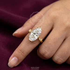 May include: A gold ring featuring a large, marquise-cut diamond as the centerpiece, flanked by smaller, round diamonds. The ring is set on a finger, showcasing the intricate design and sparkle. The ring is from NIZA GEMS.