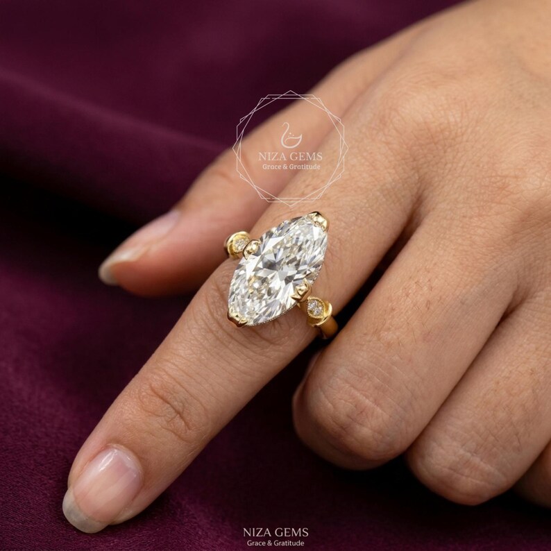 May include: A gold ring featuring a large, marquise-cut diamond as the centerpiece, flanked by smaller, round diamonds. The ring is set on a finger, showcasing the intricate design and sparkle. The ring is from NIZA GEMS.