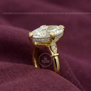 May include: A gold ring with a large, oval-cut diamond as the centerpiece. The diamond is surrounded by smaller stones and set in a gold band. The ring is on a dark purple fabric. The ring has the text "NIZA GEMS" on it.