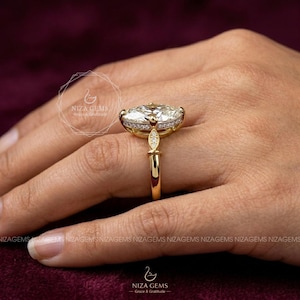 May include: A gold engagement ring with a large oval-cut diamond center stone, surrounded by smaller diamonds. The ring has a delicate band and intricate detailing. The ring is displayed on a hand, with the brand name "NIZA GEMS" visible.