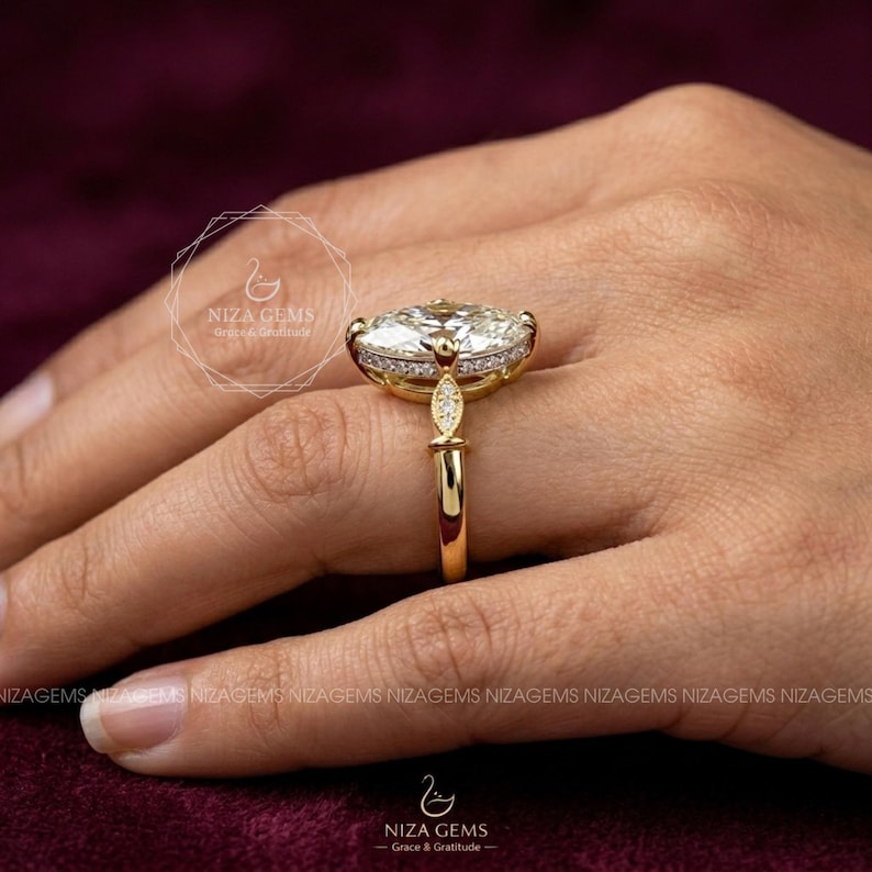 May include: A gold engagement ring with a large oval-cut diamond center stone, surrounded by smaller diamonds. The ring has a delicate band and intricate detailing. The ring is displayed on a hand, with the brand name "NIZA GEMS" visible.