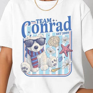 May include: White t-shirt featuring a cartoon polar bear wearing sunglasses and a scarf, with the text "Team Conrad" and "Est 2009". The bear is surrounded by seashells, starfish, and flowers, all within a blue and red frame.