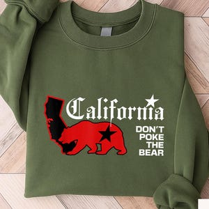 May include: An olive green sweatshirt with the word "California" in white script, a red bear graphic, and the text "DON'T POKE THE BEAR". The sweatshirt is folded on a light wood surface.
