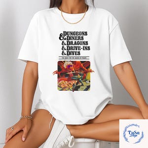 May include: White t-shirt featuring the text "DUNGEONS & DINERS & DRAGONS & DRIVE-INS & DIVES" above a colorful graphic. The graphic depicts a dragon and a knight. The text "THE QUEST FOR THE SAUCES OF POWER" is below the graphic.