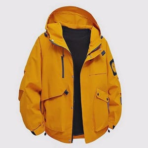 Op de afbeelding: Mosterdgele jas met capuchon en zwarte binnenkant. De jas heeft een ritssluiting aan de voorkant, meerdere zakken en een kleine patch op de bovenarm. De capuchon is bevestigd en de jas heeft een casual, sportieve stijl.