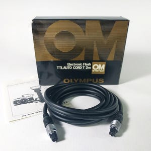 May include: Black Olympus TTLAUTO cord, 6.5 feet long, with silver connectors, and a black box with gold lettering. The box reads "OM SYSTEM" and "OLYMPUS". A manual with a camera illustration is also present.