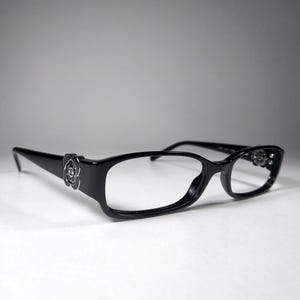 May include: Black rectangular eyeglasses featuring a glossy finish and a silver-toned camellia flower emblem on each temple. The frames have a classic shape with thin arms and integrated nose pads, providing a clean and polished look for daily wear.