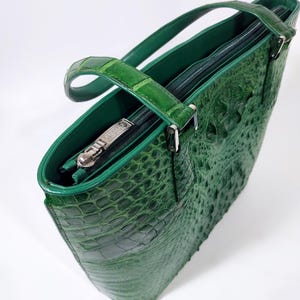 Pu&ograve; includere: Una borsa tote in pelle verde con una texture a pelle di coccodrillo. La borsa ha un manico superiore, una chiusura con cerniera e un tirazip argentato. Anche l'interno &egrave; verde. La borsa &egrave; realizzata in un materiale resistente.