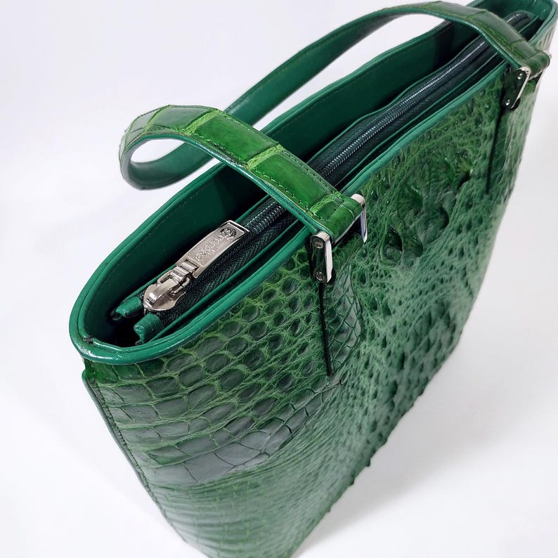 Pu&ograve; includere: Una borsa tote in pelle verde con una texture a pelle di coccodrillo. La borsa ha un manico superiore, una chiusura con cerniera e un tirazip argentato. Anche l'interno &egrave; verde. La borsa &egrave; realizzata in un materiale resistente.