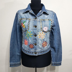 May include: A light blue denim jacket with colorful embroidered floral designs. The jacket features a classic collar, button-down front, and decorative metal studs along the pockets and hem. The floral embroidery includes shades of pink, yellow, green, and orange.