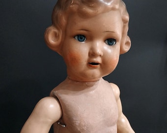 May include: Vintage doll with blue eyes, rosy cheeks, and a neutral expression. The doll has light brown hair styled in soft curls. The doll's body is a light pink color, and the arms are jointed.