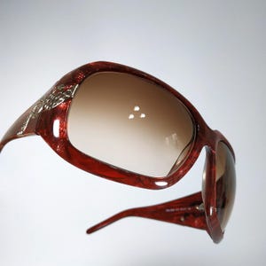 Pode incluir: Óculos de sol grandes com armação de tartaruga marmorizada em vermelho escuro e lentes castanhas degradé. As hastes apresentam um detalhe metálico prateado com cristais transparentes incrustados na lateral da armação.