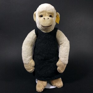 Puede incluir: Un muñeco de mono de peluche con cara, manos y pies de color crema, vestido con un vestido gris oscuro. El muñeco tiene una etiqueta amarilla sujeta a la oreja y está de pie sobre una base blanca.