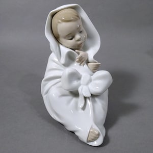Puede incluir: Figura de porcelana de un bebé envuelto en una manta blanca con un gran lazo. La cara y las manos del bebé son visibles, con una expresión serena. La figura es de color blanco suave.