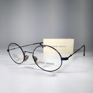 May include: Black round-frame eyeglasses with the Giorgio Armani logo printed on the lens. The glasses have a thin, dark frame and a small, light-colored nose pad. A small, rectangular box is in the background.