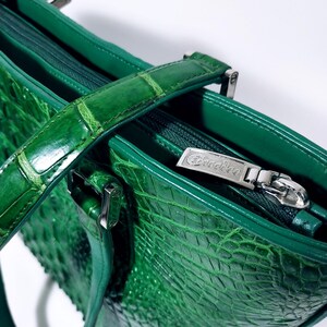 Pu&ograve; includere: Primo piano di una borsa in pelle verde con una texture a pelle di coccodrillo. La borsa presenta un manico superiore, una chiusura con cerniera e un tirazip argentato. L'interno &egrave; visibile e la borsa sembra nuova.