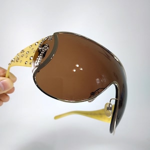 以下が含まれることがあります： ブラウンの連続したレンズと金属フレームを備えたオーバーサイズのシールドサングラス。テンプルはライトイエローで、小さな金属製のスタッドとヒンジ付近の目立つシルバーカラーのロゴエンブレムで装飾されています。