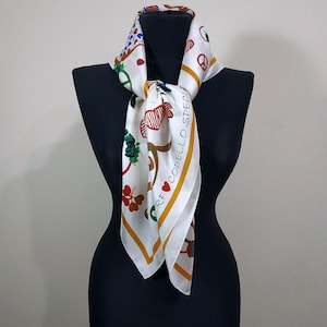 May include: White silk scarf with a colorful pattern of hearts, flowers, and other designs. The scarf has a gold border and the text "RF CODELLO SPECIAL". The scarf is draped over a black mannequin.