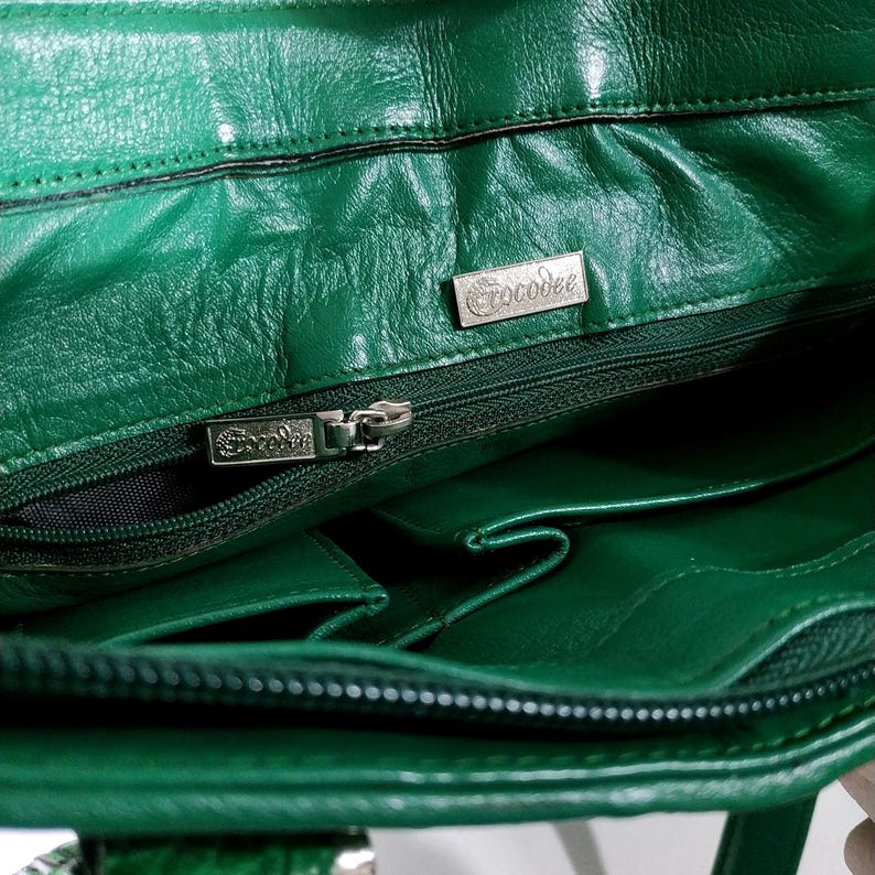 Pu&ograve; includere: Primo piano dell'interno di una borsa in pelle verde. La borsa presenta pi&ugrave; scomparti, una chiusura con cerniera e un logo in metallo argentato con la scritta "Crocodee". La pelle ha un aspetto strutturato.