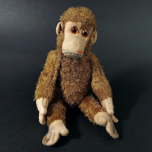 Op de afbeelding: Een vintage pluchen aap met bruine vacht, een beige gezicht, handen en voeten. De aap heeft grote, ronde, oranje ogen en een kleine rode mond. Hij zit met uitgestrekte benen en armen langs zijn zij.