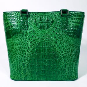 Pu&ograve; includere: Una borsa tote verde brillante con una texture di pelle di coccodrillo. La borsa ha una forma rettangolare con un design strutturato, cuciture visibili e hardware argentato sui manici. La borsa &egrave; realizzata in un materiale resistente.