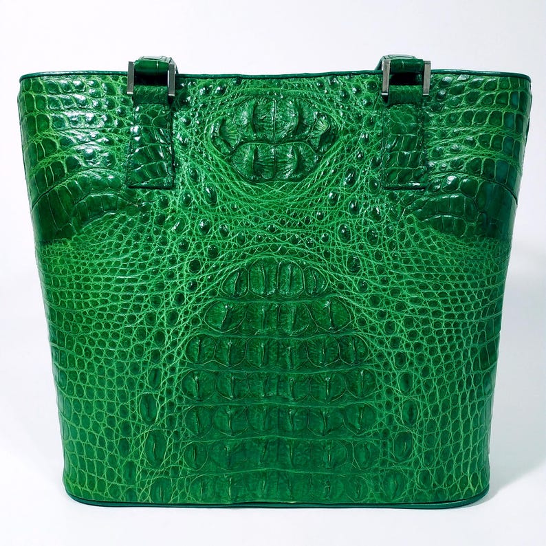 Pu&ograve; includere: Una borsa tote verde brillante con una texture di pelle di coccodrillo. La borsa ha una forma rettangolare con un design strutturato, cuciture visibili e hardware argentato sui manici. La borsa &egrave; realizzata in un materiale resistente.