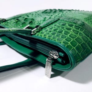 Pu&ograve; includere: Una borsa in pelle verde con una texture a pelle di coccodrillo. La borsa presenta una chiusura con cerniera con un tirante argentato e una fibbia argentata. La borsa ha un manico e un design strutturato.