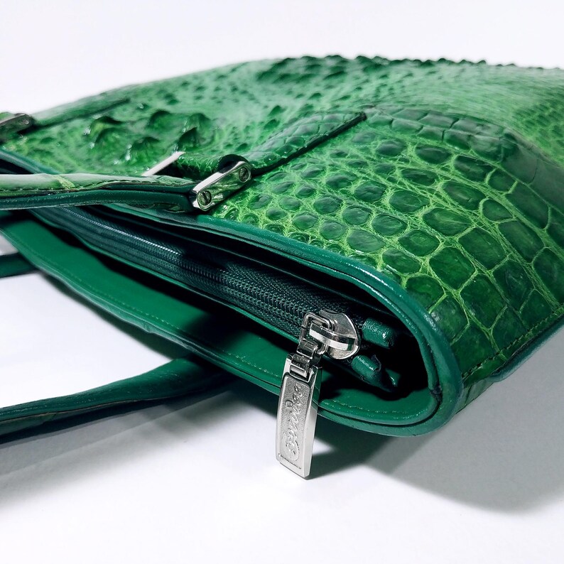 Pu&ograve; includere: Una borsa in pelle verde con una texture a pelle di coccodrillo. La borsa presenta una chiusura con cerniera con un tirante argentato e una fibbia argentata. La borsa ha un manico e un design strutturato.