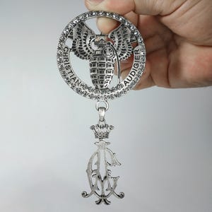 Puede incluir: Un colgante de metal plateado con un marco circular incrustado con pedrería transparente. El centro muestra un gráfico de granada alada con el texto CHRISTIAN AUDIGIER. Un dije colgante debajo presenta una corona sobre las letras CA.