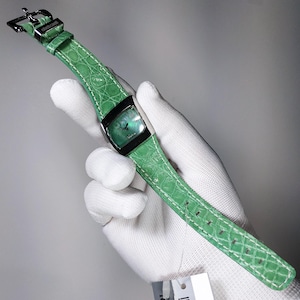Puede incluir: Un reloj de pulsera verde con una esfera rectangular y una correa de cuero verde a juego. El reloj tiene un bisel negro y una hebilla plateada. La correa presenta una textura de piel de cocodrilo y costuras blancas.