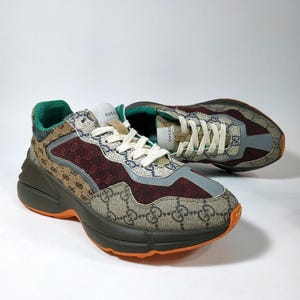 May include: A pair of multi-colored sneakers with a brown and beige GG monogram pattern. The sneakers have burgundy and gray accents, green trim, and orange soles. White laces and the Gucci logo are visible.